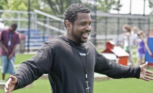 A smiling man on a sports pitch opens his arms wide.